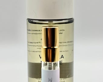 May include: A clear glass bottle with a white cap and a smaller perfume bottle in front. The smaller bottle has a gold cap and the words "VANILLA SMOKE" printed on it. The background is white.