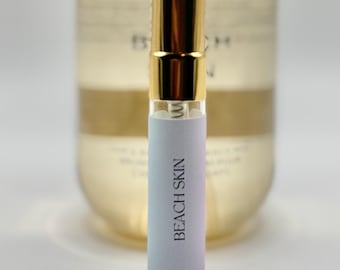 May include: A small perfume vial with a gold cap and a light blue label that reads "BEACH SKIN" in black text. The vial is in front of a larger, blurred bottle with a gold cap.