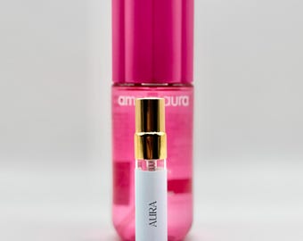 May include: A small perfume sample labeled "AURA" with a gold cap and a larger pink bottle in the background. The sample is in a clear glass vial with a white label. The larger bottle has a pink cap and a pink liquid inside.