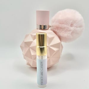 May include: A small, clear glass bottle with a gold cap and a white label that says "Sweet Like Candy". The bottle is sitting in front of a larger, pink bottle with a fluffy pink pom-pom on the top.