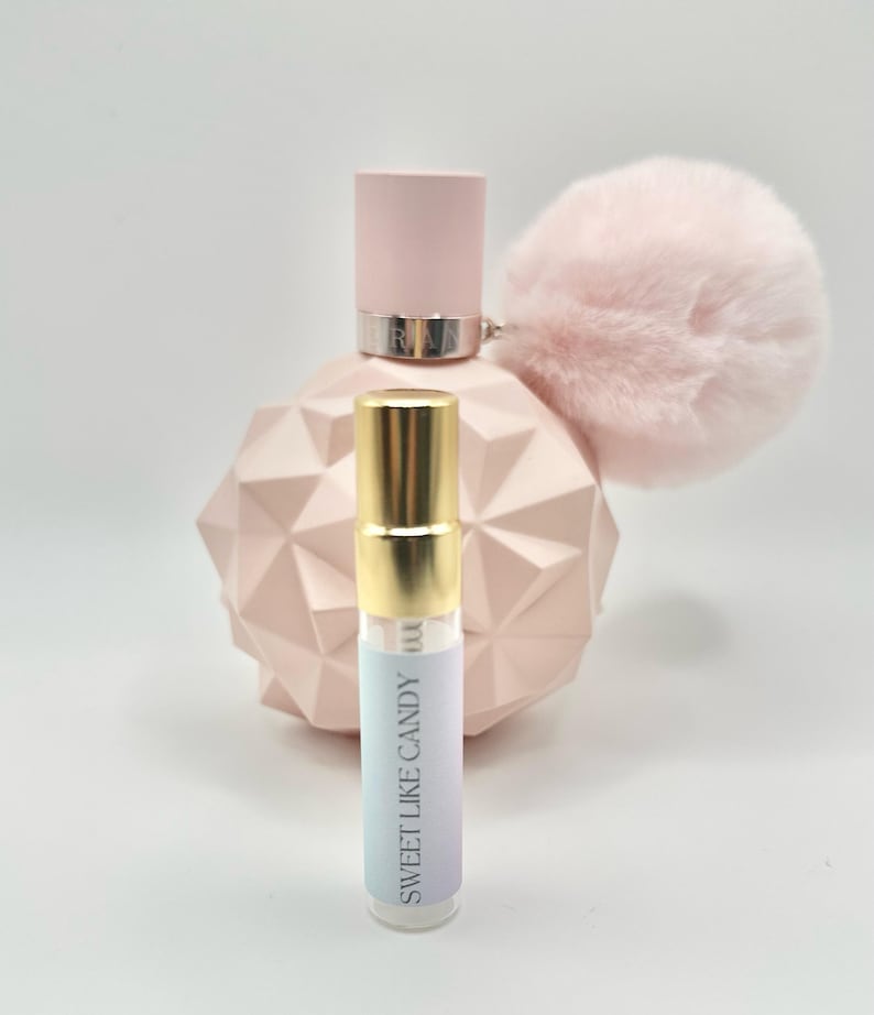 May include: A small, clear glass bottle with a gold cap and a white label that says "Sweet Like Candy". The bottle is sitting in front of a larger, pink bottle with a fluffy pink pom-pom on the top.