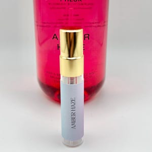 May include: A small clear glass bottle with a gold cap and a white label that says "Amber Haze". The bottle is in front of a larger pink bottle with a white label that says "Amber Haze".