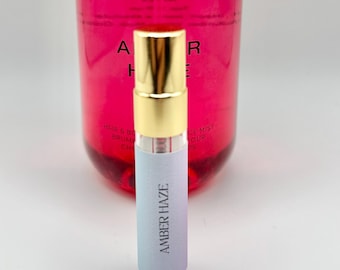 May include: A small clear glass bottle with a gold cap and a white label that says "Amber Haze". The bottle is in front of a larger pink bottle with a white label that says "Amber Haze".