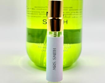 May include: A small perfume vial with a gold cap and a white label that reads "MRS. SMITH." The vial is in front of a large, green bottle with text. The background is a soft white.