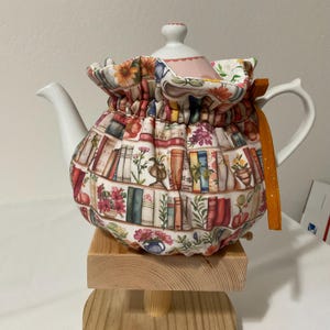 May include: White ceramic teapot with a decorative fabric cozy. The cozy features a colorful print of books, flowers, and vases. An orange ribbon is tied to the handle. The teapot is sitting on a small wooden stand.