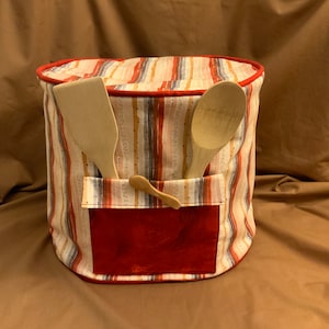 May include: A red, white, and brown striped fabric cover for a slow cooker with a red pocket and two wooden spoons sticking out of the top.