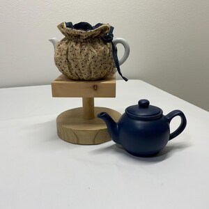 May include: A white teapot with a floral patterned cozy and a navy blue teapot. The cozy is on a wooden stand. The navy teapot is on a white surface.