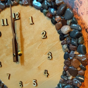 May include: A round wooden clock with a light brown face and a border of various-sized, multi-colored stones. The clock hands and numerals are a darker brown. The numerals are in a simple, sans-serif font.