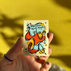 Könnte beinhalten: Ein rechteckiger Aufkleber mit den Worten "PROTECT YOUR PEACE" in schwarzer Schrift. Der Aufkleber zeigt eine farbenfrohe Illustration einer abstrakten Figur in Orange, Blau und Gelb. Der Aufkleber wird in einer Hand mit blauem Nagellack und einem blauen Perlenring gehalten.