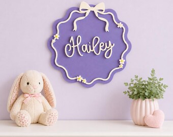 May include: A purple scalloped wall sign featuring the name Hailey in white script lettering. The sign is decorated with a white bow at the top and small white daisy accents along the border, creating a decorative piece for a wall display.