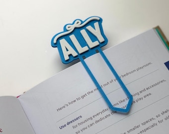 May include: A blue and white bookmark with the name "ALLY" in white block letters. The bookmark is attached to the pages of an open book. The bookmark has a bow design at the top.