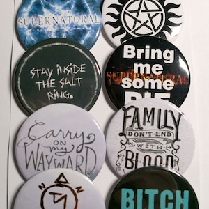 Puede incluir: Nueve botones redondos con temática de Supernatural con varios diseños y textos. Los botones presentan combinaciones de colores negro y blanco, azul y blanco, y negro y gris. El texto en los botones incluye "Supernatural", "Quédate dentro del círculo de sal", "Sigue mi camino rebelde", "Tráeme algo de pastel", "La familia no termina con sangre", y "Perra idiota".