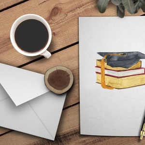 May include: A white envelope, a cup of coffee, a wooden coaster, and a white card with a watercolor illustration of a graduation cap on top of a stack of books.