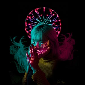 May include: A person with pink and blue hair is wearing neon yellow makeup on their face. The makeup is in the form of Korean characters. The person is wearing a yellow sweater and is holding their hands in front of their face. A pink and blue neon light is behind the person.