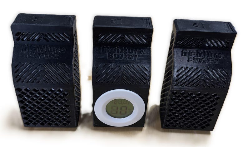 K&ouml;nnte beinhalten: Drei schwarze Kunststoff-Feuchtigkeitsabsorber mit dem Aufdruck "Moisture Buster". Der mittlere Absorber hat ein wei&szlig;es digitales Hygrometer mit einer Anzeige von 88%.