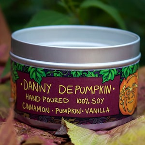 May include: A silver tin candle with a purple label that reads "Danny DePumpkin: Hand Poured 100% Soy Cinnamon - Pumpkin - Vanilla". The label features a cartoon pumpkin with glasses.