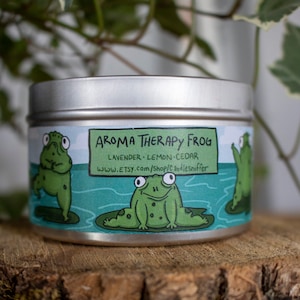 May include: A silver tin candle with a label featuring cartoon frogs on lily pads. The label reads "Aroma Therapy Frog" with scent notes of lavender, lemon, and cedar. The Etsy shop URL is also included. The candle sits on a wooden surface.