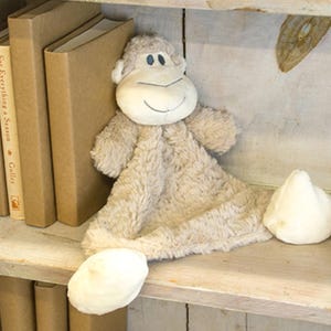 May include: A plush, beige monkey toy with a smiling face and floppy limbs rests on a wooden shelf. The shelf also holds several hardcover books and a small wooden crate. Three decorative candles sit above.