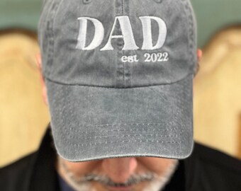 Op de afbeelding: Een grijze katoenen baseballpet met witte geborduurde tekst die "DAD est 2022" luidt.