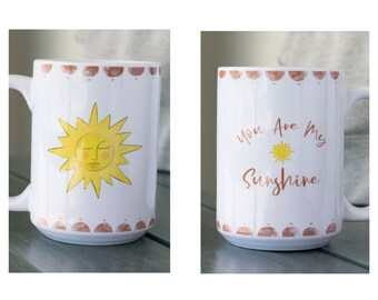 May include: White ceramic mug featuring a yellow sun illustration with a sleepy face and the text "You Are My Sunshine." The mug has a decorative border of brown shapes. A cheerful and bright design for a coffee or tea cup.