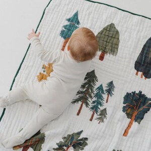 May include: White baby blanket with a green border, featuring a repeating pattern of illustrated trees in various colors. The blanket is made of a soft, textured fabric. A baby in a white onesie is lying on the blanket.