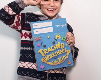 Pode incluir: Uma criança segura um livro azul "Tracing Worksheet for Kids" com 71 páginas. O livro tem ilustrações e texto coloridos. A criança usa um suéter azul marinho e cinza com um padrão vermelho e branco e jeans.