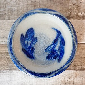 May include: A handmade ceramic bowl with a white interior and a blue, textured exterior. The bowl features a blue floral design in the center and a wavy blue pattern around the rim. The bowl is a decorative piece for the kitchen or dining room.
