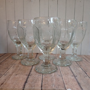 May include: Set of six clear textured glass goblets. Each goblet has a textured bowl, a stem, and a textured base. The goblets are arranged on a light brown wooden surface. The glass has a vintage look.