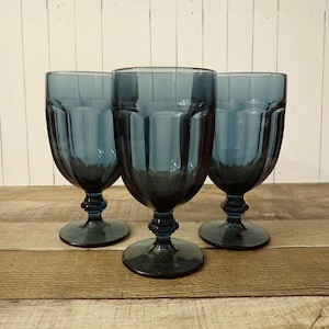 May include: Three blue-tinted glass goblets with a ribbed design. The goblets have a rounded bowl, a detailed stem, and a circular base. They are arranged on a wooden surface against a white-painted wood background.