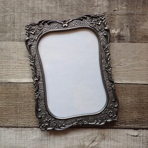 May include: A rectangular picture frame with a metallic silver finish, featuring an ornate border decorated with raised floral motifs and a textured, stippled background. The frame surrounds a plain white rectangular insert with rounded corners.