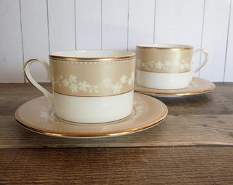 May include: Two matching teacups and saucers. The cups have a tan band with white floral designs and a white base. The saucers and rims of the cups are trimmed with gold. The set is on a wooden surface.