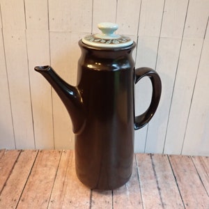 May include: A brown ceramic coffee pot with a light blue lid and a brown and white floral design. The pot has a tall, slender shape and a curved handle.