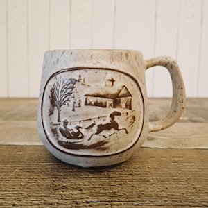 May include: A speckled beige ceramic mug with a brown embossed rural scene featuring a barn, tree, and horse-drawn sleigh. The mug has a large handle and a rustic, vintage aesthetic.