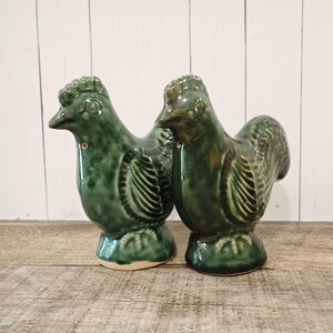 May include: Two ceramic rooster figurines in a dark green glaze. The roosters have detailed feather and comb designs. They are standing on a wooden surface with a white wooden background.