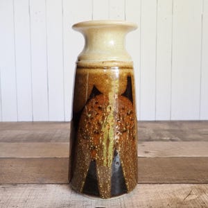 May include: A tall, ceramic vase with a flared top and a textured, geometric design. The vase features a light beige top and a body with brown, black, and green accents. The vase is approximately 10 inches tall.