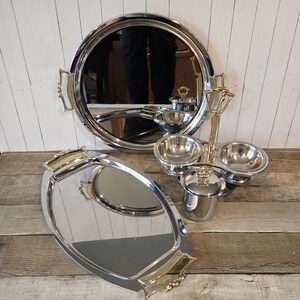 May include: A set of polished silver-tone metal serving pieces including a large round tray, an oval tray, and a condiment server with three bowls and a central handle. Each piece features decorative gold-tone metal handles and accents.