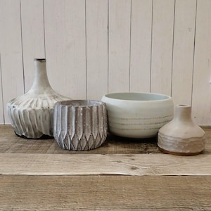 May include: Four ceramic vases in various shapes and sizes. The vases are all white with a textured finish. The vases are arranged on a wooden surface.