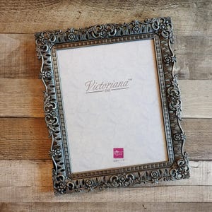 May include: A rectangular picture frame with an ornate, silver-toned metallic border featuring intricate floral and scrollwork relief patterns. The center insert displays the text Victoriana and a small pink Russ logo with the text size 8 x 10 inches.