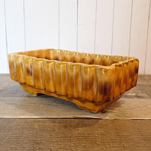 May include: A rectangular, vintage-style planter with a ribbed design. The planter is glazed in shades of brown and yellow, with darker accents. It sits on a wooden surface, suggesting a rustic or farmhouse aesthetic. The planter is suitable for indoor or outdoor use.