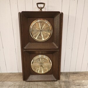 May include: A vintage wooden weather station with two brass-faced gauges. The gauges are set in a dark brown wooden frame with a brass handle at the top. The gauges display temperature and pressure readings.