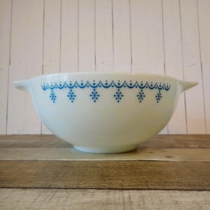 May include: White mixing bowl with a blue snowflake and dotted pattern around the rim. The bowl has two handles and sits on a wooden surface. This vintage kitchenware is ideal for baking and food preparation.