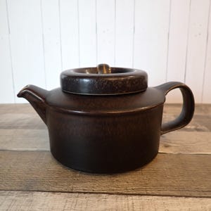 May include: A white ceramic mug with a black handle and the text "But First, Coffee" in a bold, black font. The mug is set against a blurred background, suggesting a cozy setting.