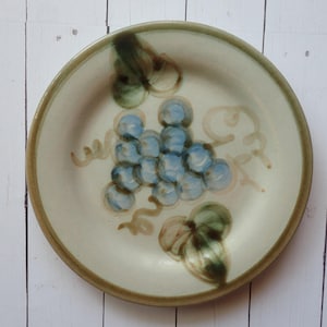May include: A ceramic plate with a hand-painted grape design. The plate features a beige background with a brown rim and a cluster of blue grapes with green leaves. The design is simple and rustic, suitable for serving food.