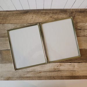 May include: A hinged, gold-colored picture frame with two square openings. The frame has a decorative beaded edge and is set against a wood-grain background. The frame is empty, ready for photos or artwork.