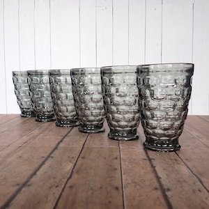 May include: Six gray glass tumblers with a textured, honeycomb-like design. The glasses are arranged in a row on a wooden surface.
