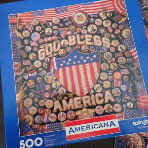 Może przedstawiać: 500-elementowa układanka Americana o tematyce patriotycznej. Układanka przedstawia wz&oacute;r flagi amerykańskiej, słowa "GOD BLESS AMERICA" i r&oacute;żne patriotyczne obrazy. Pudełko jest niebieskie i ma logo "Americana".