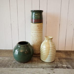 May include: Three ceramic vessels including a round dark green vase, a tall cylindrical vase with a cream speckled base and a dark green and brown top, and a smaller cream-colored vase with a ribbed texture and a wide neck opening.