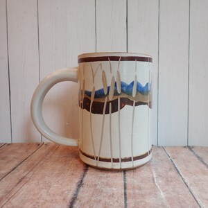 May include: White ceramic mug with a brown, blue, and gold design of tall grass. The design features a brown band with a blue and gold stripe.