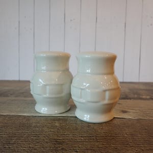 May include: A pair of off-white ceramic salt and pepper shakers featuring a rounded, bulbous shape with a textured, basket-weave pattern around the center. Each shaker has a smooth, flared top rim and a matching flared base for stability.
