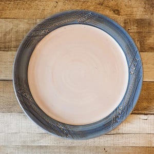 May include: A handmade ceramic plate with a light pink center and a blue rim. The rim features a textured design with repeating patterns. The plate is round and sits on a wooden surface, showcasing its handcrafted details.